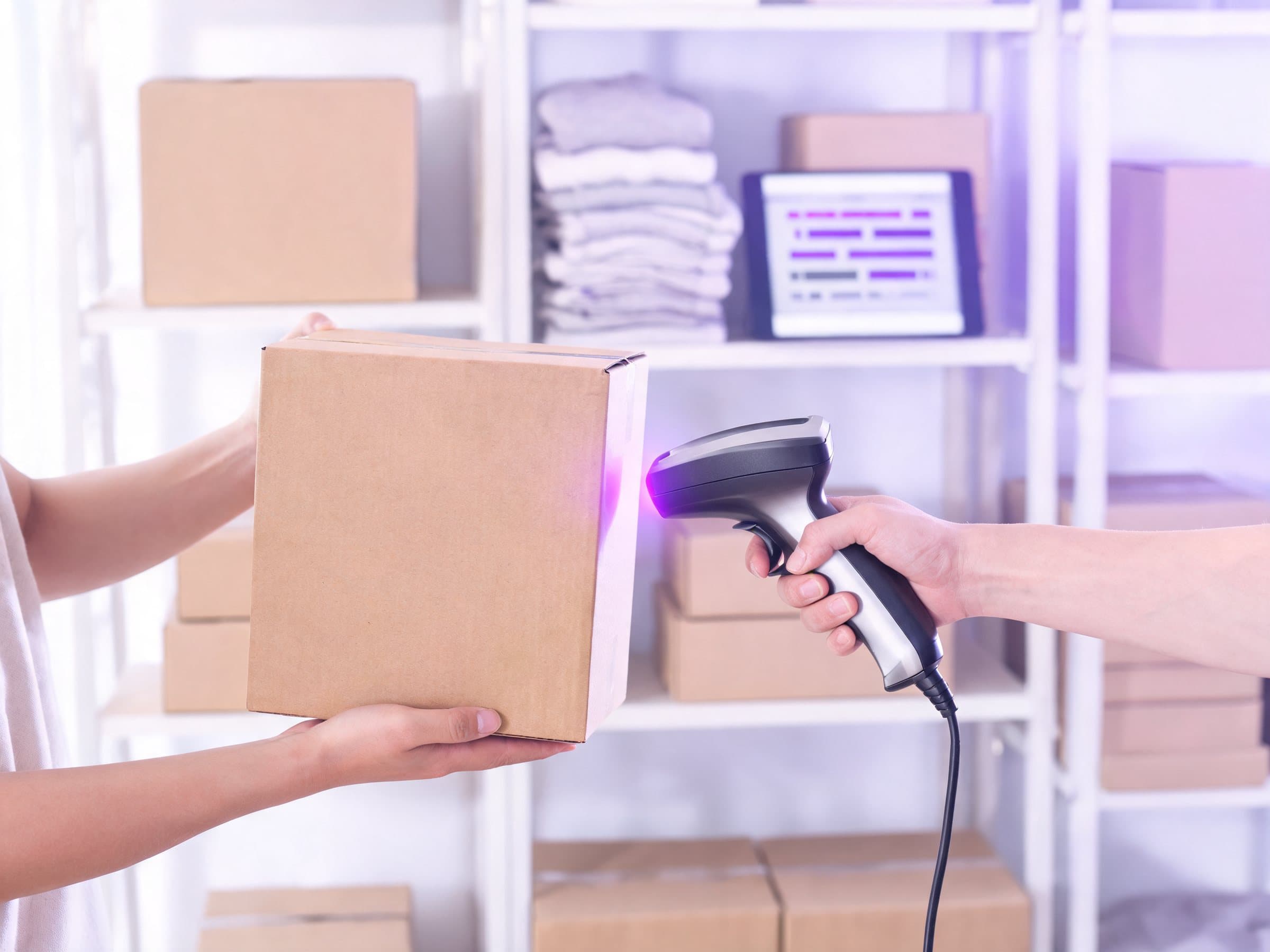 A handheld scanner reading a cardboard box in a Dubai boutique’s stockroom, shelves of boxes and folded clothes behind