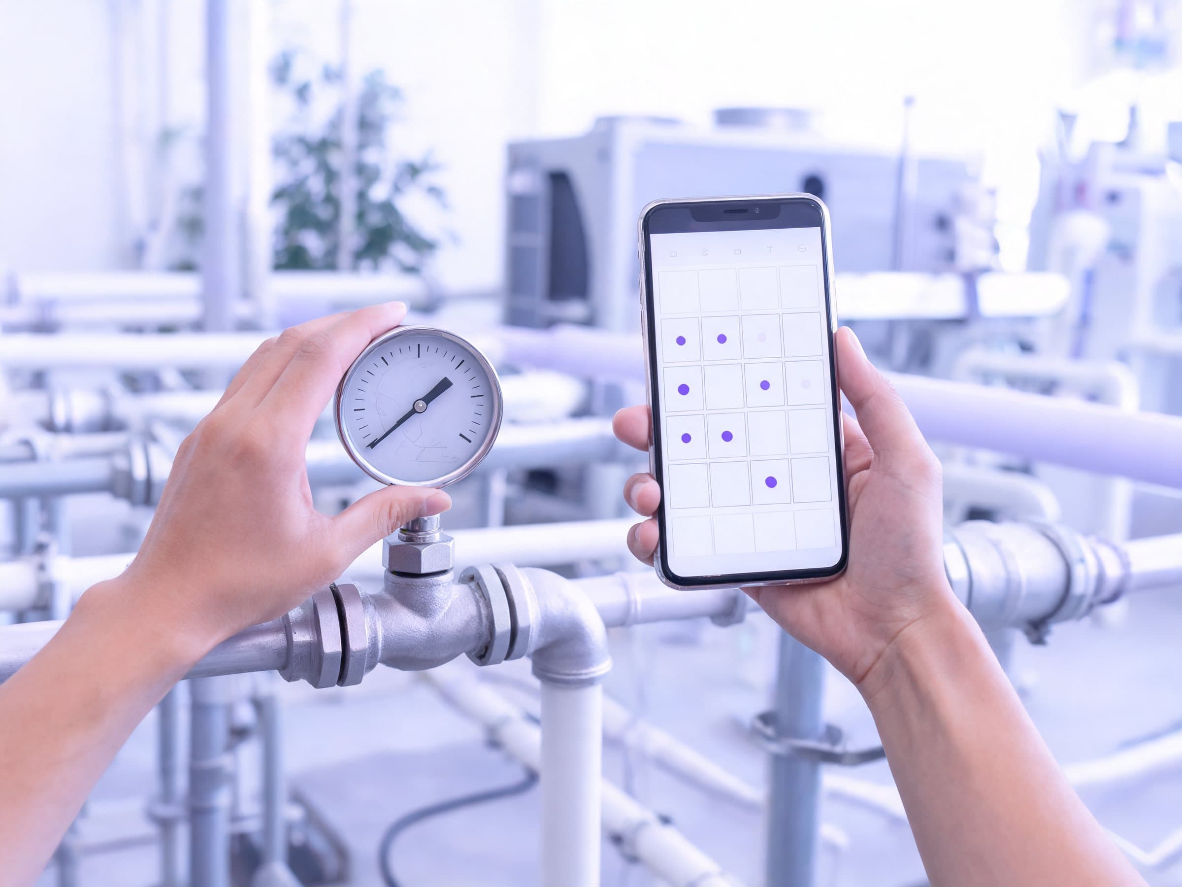 One hand checking a pressure gauge on pipework in a Dubai plant room, the other holding a phone with a monthly calendar